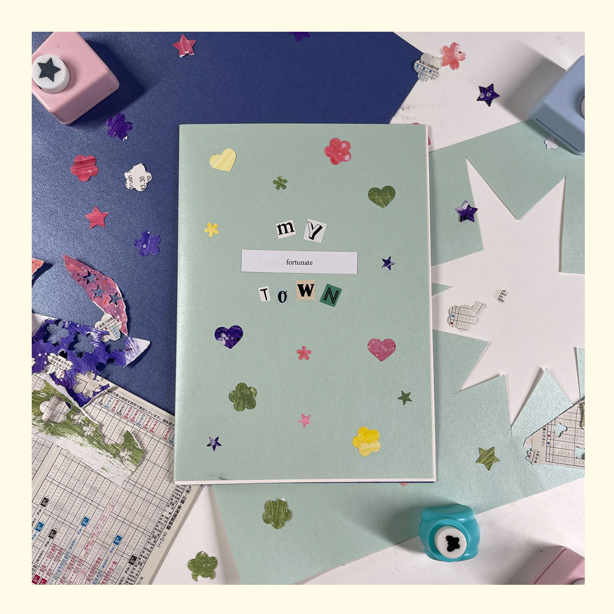 the cover page of a zine called 'my fortunate town' surrounded by the materials it is made with (pearl paper, painted collage paper, paper cut outs, train timetables)